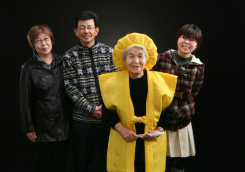 米寿祝。黄色のちゃんちゃんこの＜母と祖母＞を囲む子ども夫婦とお孫さんの記念写真。